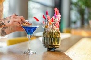 A martini glass with a blue liquid on a wooden table next to a decorative vase with various flowers. - Novotel Muenchen City (Munich)