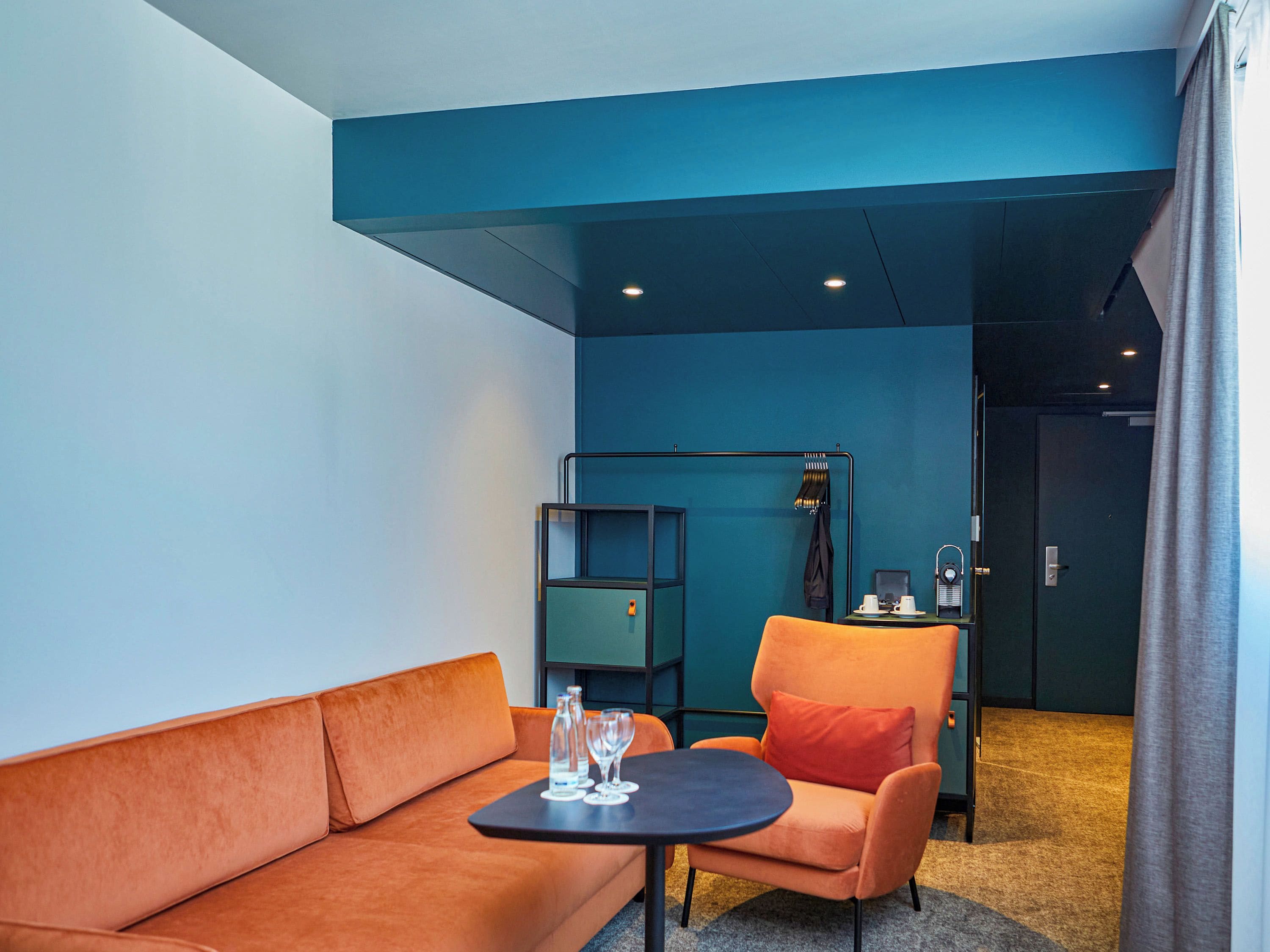 Una habitación de hotel moderna con sofá, sillón y una pequeña mesa con vasos y una bandeja.