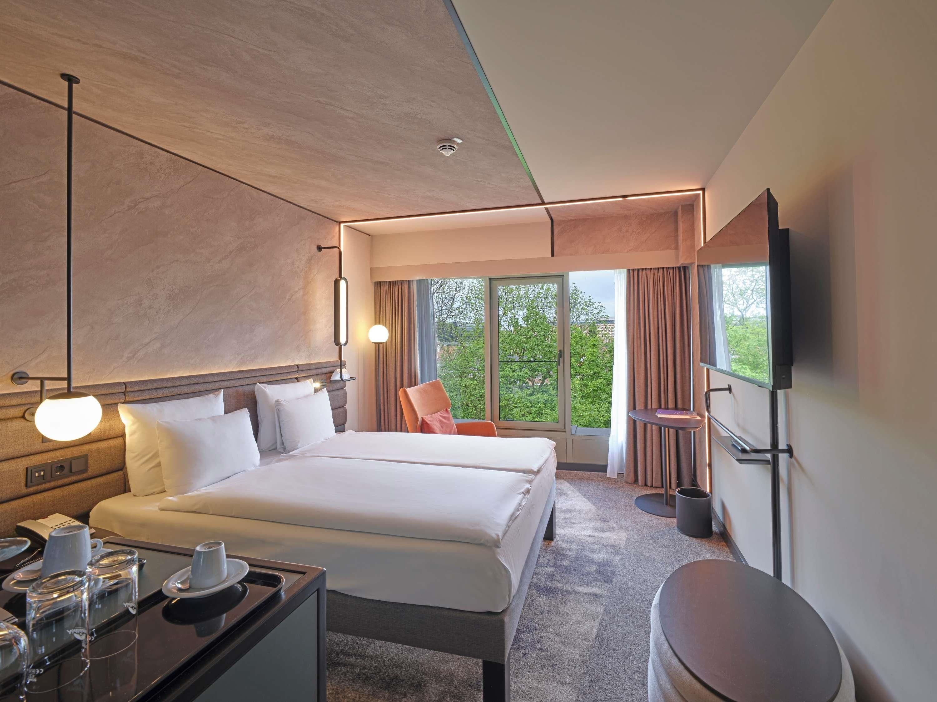 A modern hotel room with a large bed, bedside tables, a desk with a telephone, and a TV mounted on the wall.