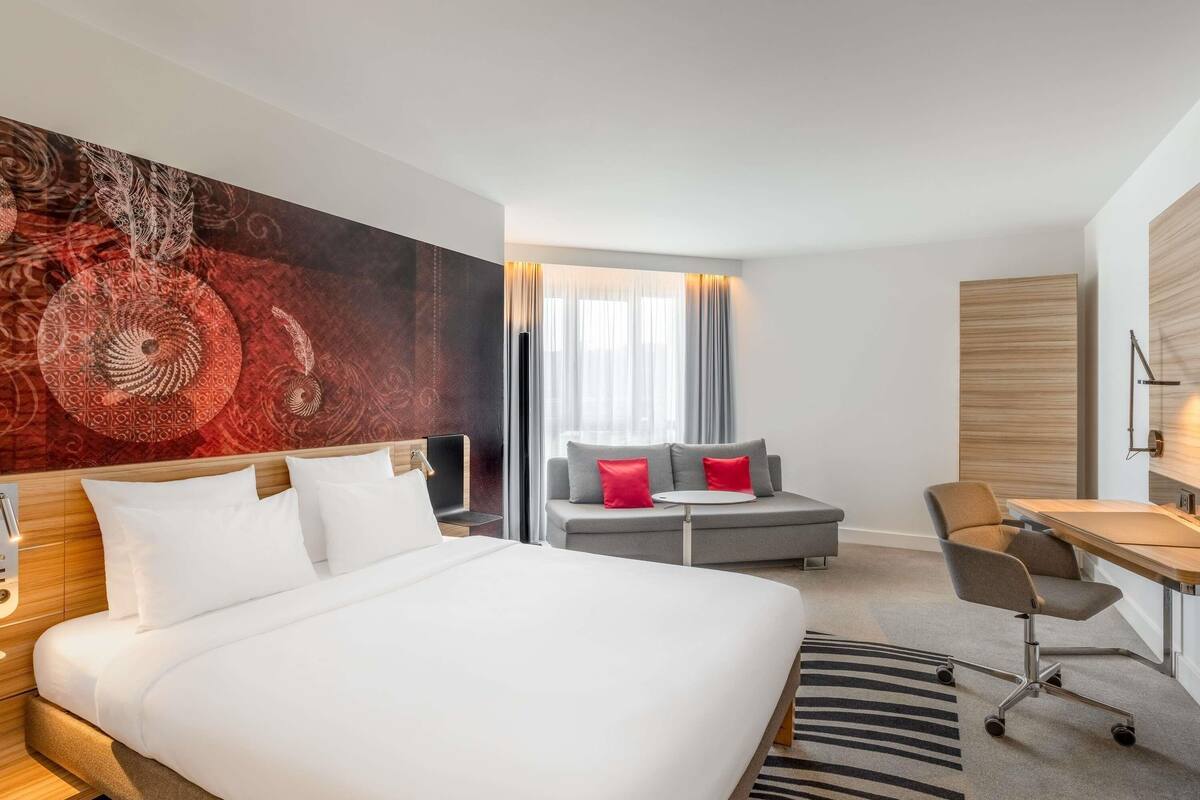 Ein Hotelzimmer mit einem großen Bett, einem grauen Sofa, einem Schreibtisch und einem großen abstrakten Wandbild.