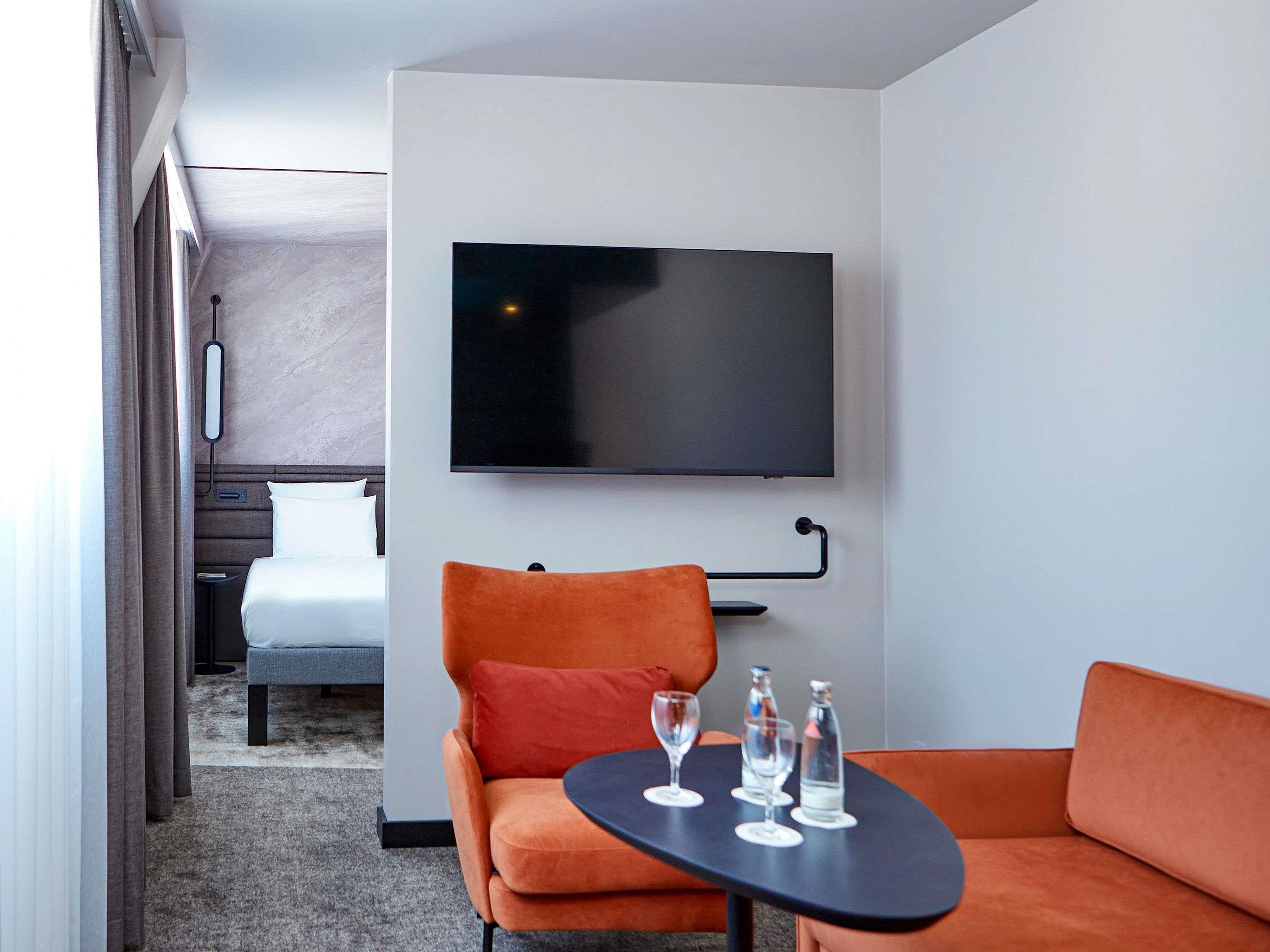 Une chambre d’hôtel moderne avec un lit, un téléviseur fixé au mur, un canapé orange, une petite table avec des verres et des bouteilles d’eau, et un sol recouvert de moquette.