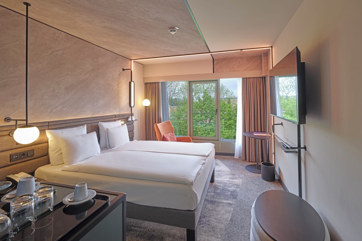A hotel room with a large bed, bedside tables, a desk with a telephone and glasses, a chair, a TV, and a window with a view of trees.
