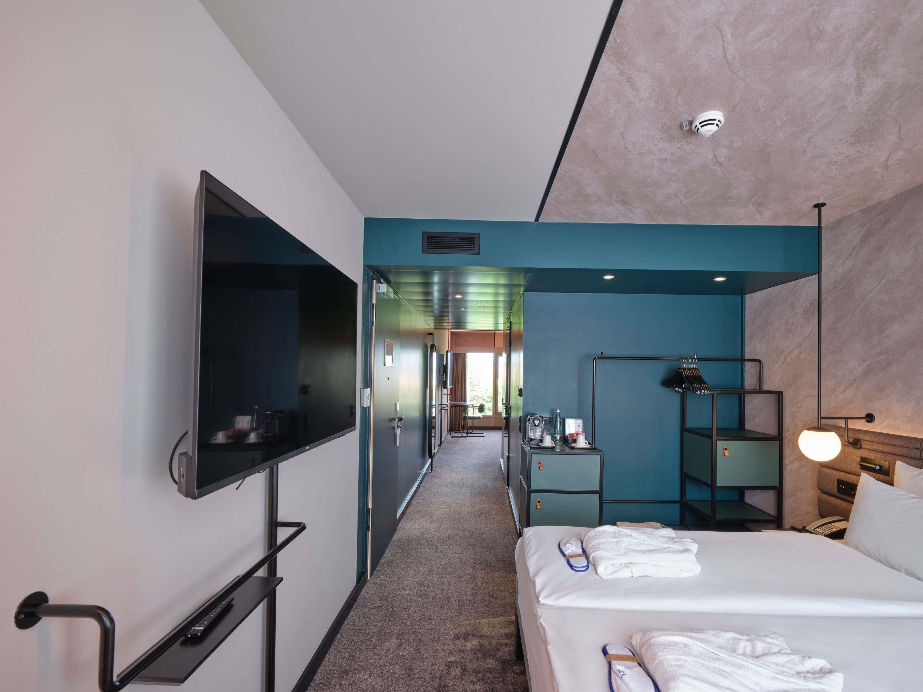 A hotel room with a flat-screen TV, a bed with white linens, a desk with a lamp, and a view of the hallway.