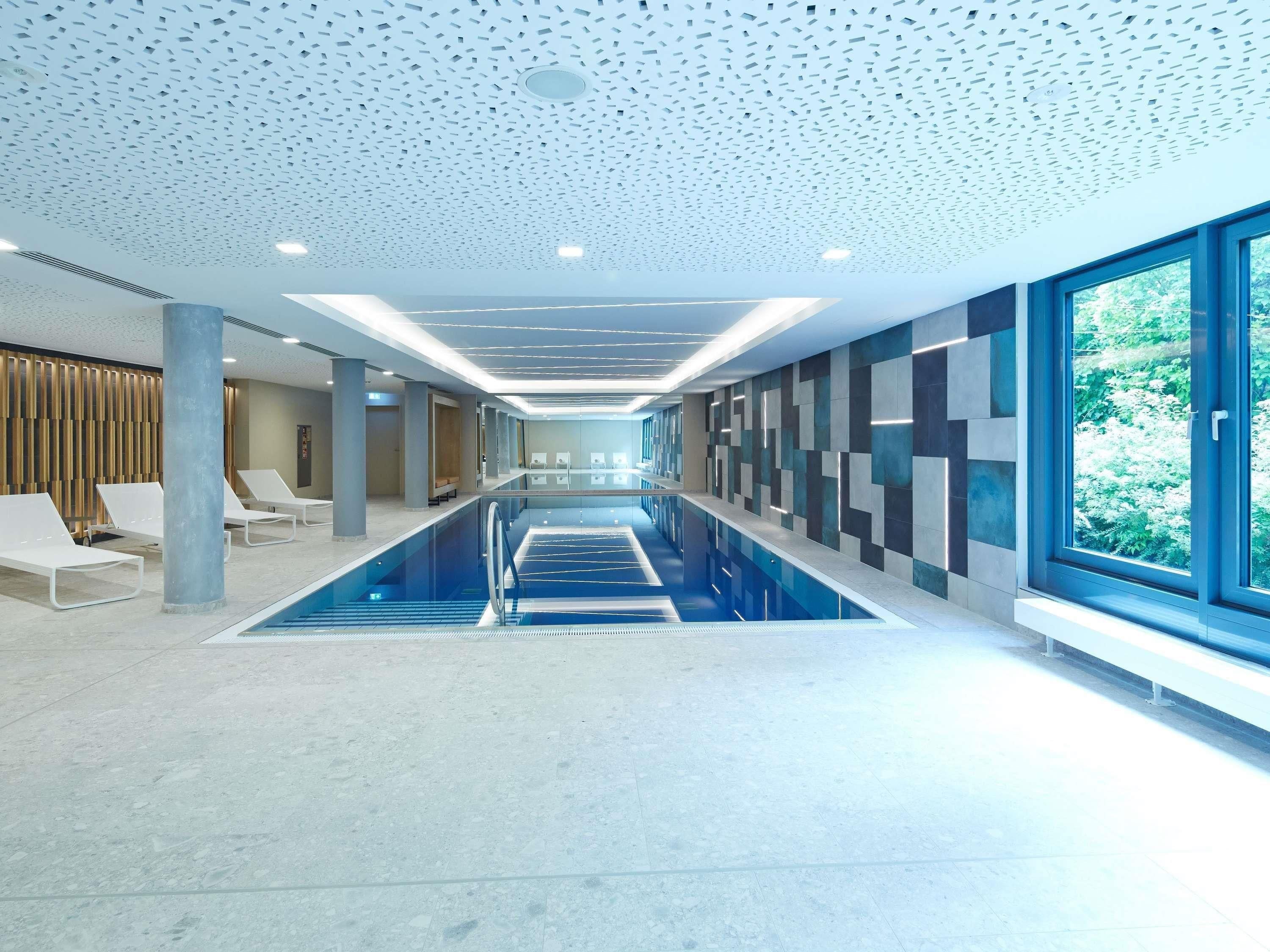 A modern indoor swimming pool with a long, narrow design, a ceiling with a geometric pattern, and a view of greenery outside.