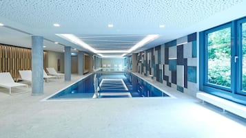 A modern indoor swimming pool with a long, narrow design, a ceiling with a geometric pattern, and a view of greenery outside.