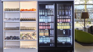 A beverage refrigerator stocked with various drinks, including water, soda, and beer, in a modern setting with a view of the outside. - Novotel Muenchen City (Munich)