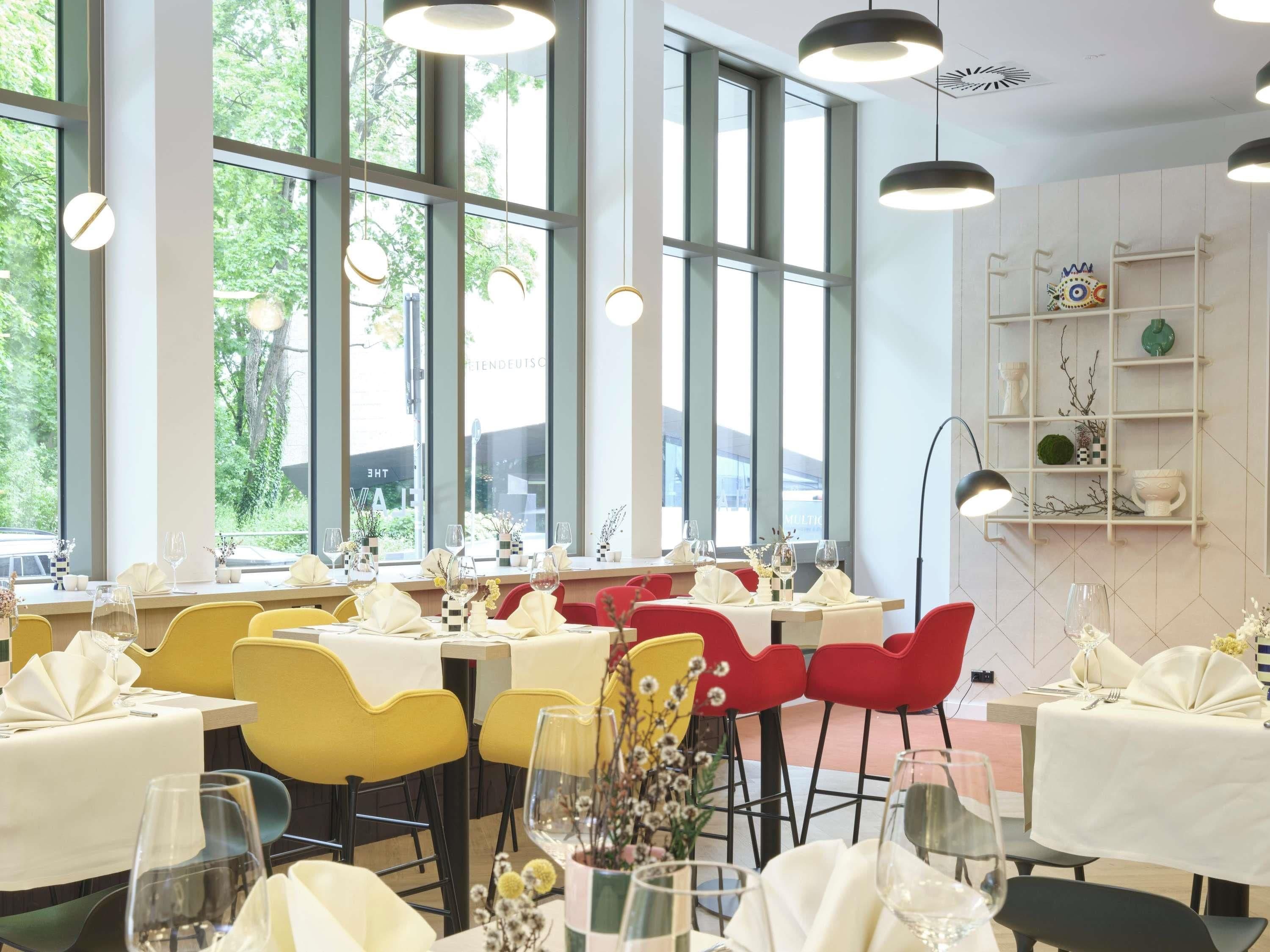 A restaurant with multiple tables, each set with white tablecloths and red chairs. Large windows allow natural light to fill the space.