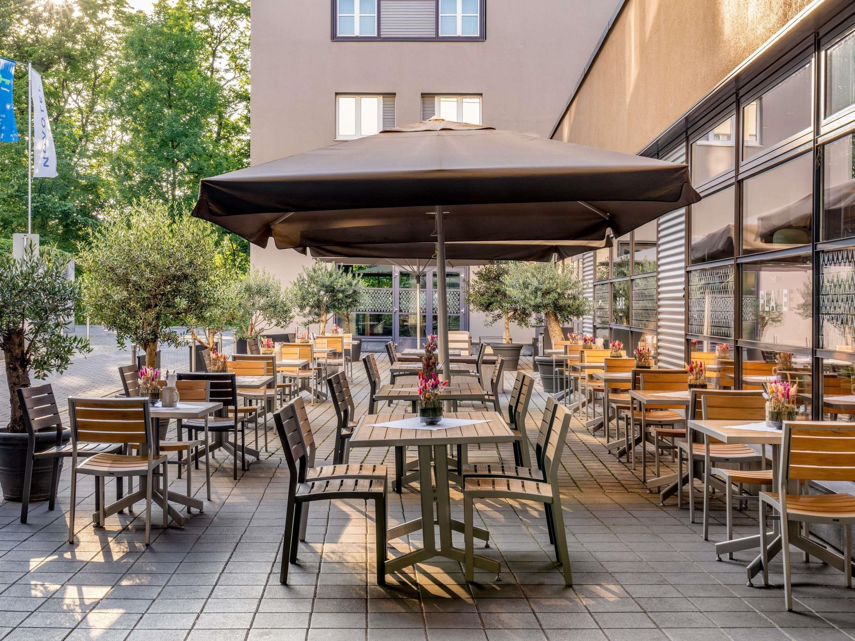 An outdoor patio with tables and chairs, surrounded by trees and a building with large windows.