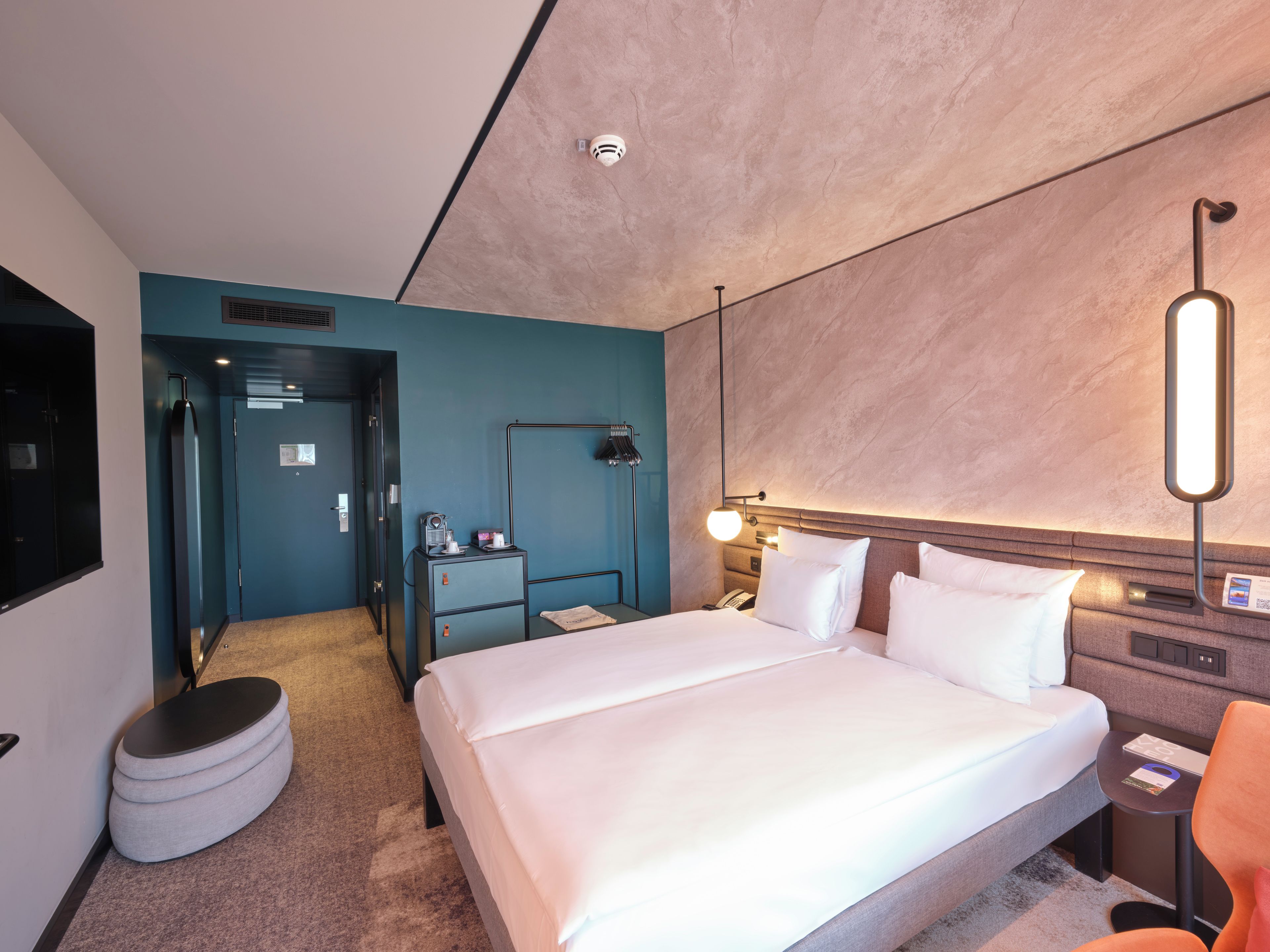 Ein modernes Hotelzimmer mit einem großen Bett, einem Schreibtisch, einem Sessel, einer Lampe und einer Minibar.