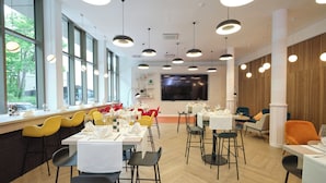 A modern restaurant with a long counter, yellow and black chairs, and a large window with a view of trees. - Novotel Muenchen City (Munich)