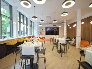 A modern restaurant with a long counter, yellow and black chairs, and a large window with a view of trees. - Novotel Muenchen City (Munich)
