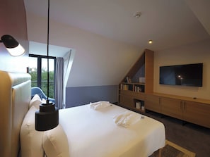 A modern hotel room with a large bed, a flat-screen TV, a built-in bookshelf, and a balcony with a view of trees. - Novotel Muenchen City (Munich)