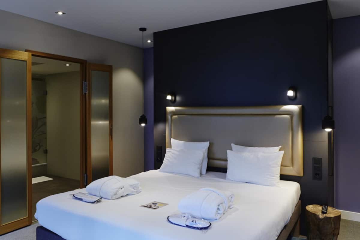 Ein modernes Hotelzimmer mit einem großen Bett, zwei Nachttischlampen, einem Holznachttisch und einem Spiegel.