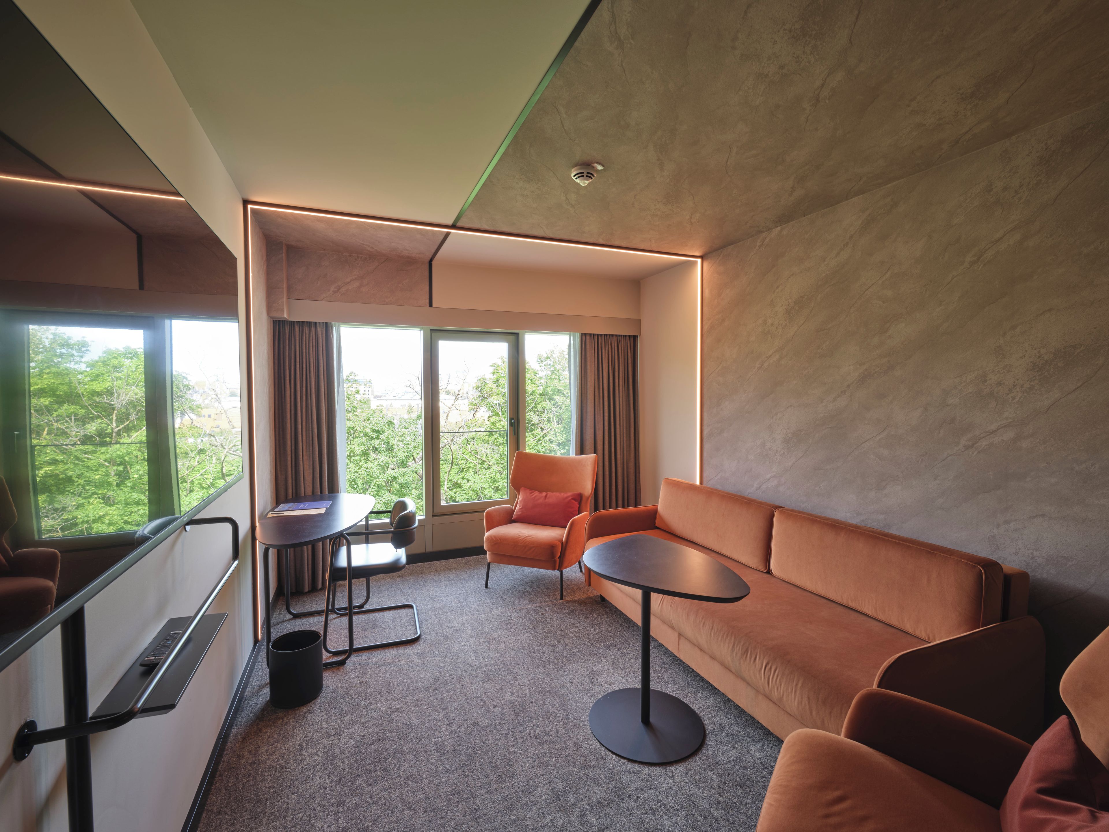 A modern hotel room with a sofa, armchairs, a small table, and a view of greenery outside.