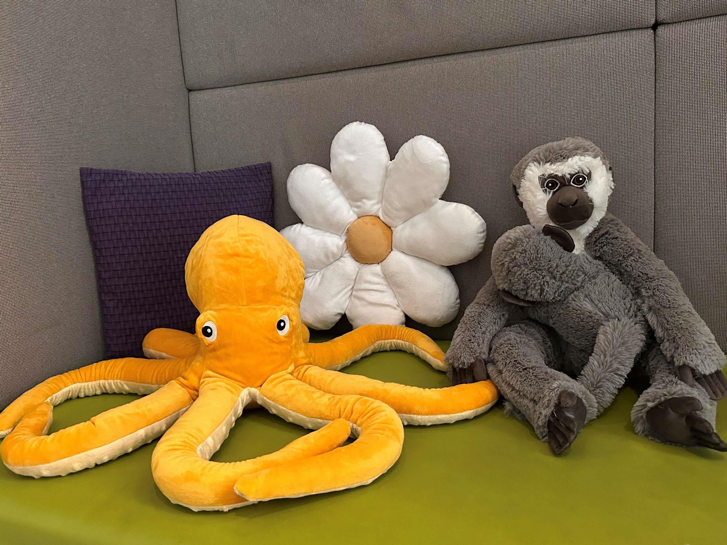 A sofa with a purple pillow, a white flower-shaped pillow, and a gray stuffed animal.
