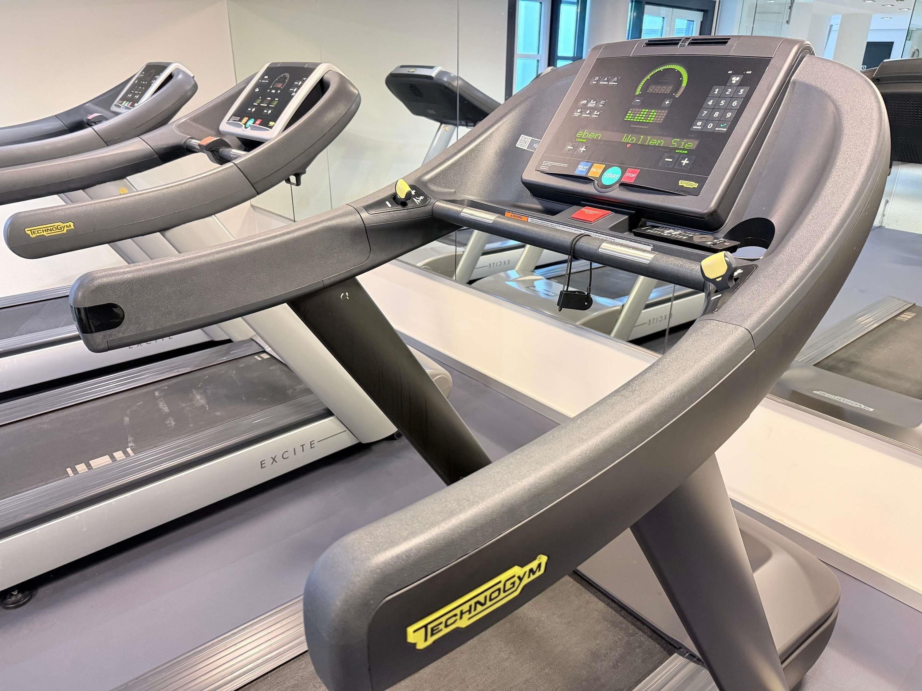 A treadmill with a digital display and control panel, positioned in a gym with other exercise equipment.