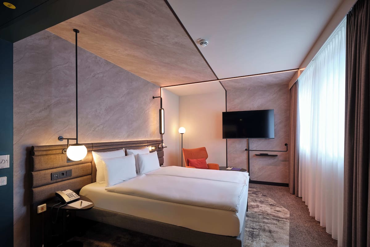 Ein modernes Hotelzimmer mit einem großen Bett, einem Nachttisch mit Telefon und Notizblock, einem an der Wand montierten Flachbildfernseher und einem großen Fenster mit Vorhängen.