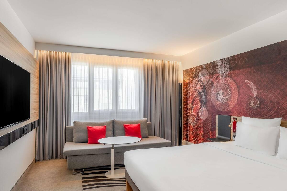 Ein Hotelzimmer mit einem Bett, einer Couch mit roten Kissen, einem kleinen runden Tisch, einem an der Wand montierten Fernseher und einem großen abstrakten Kunstwerk.