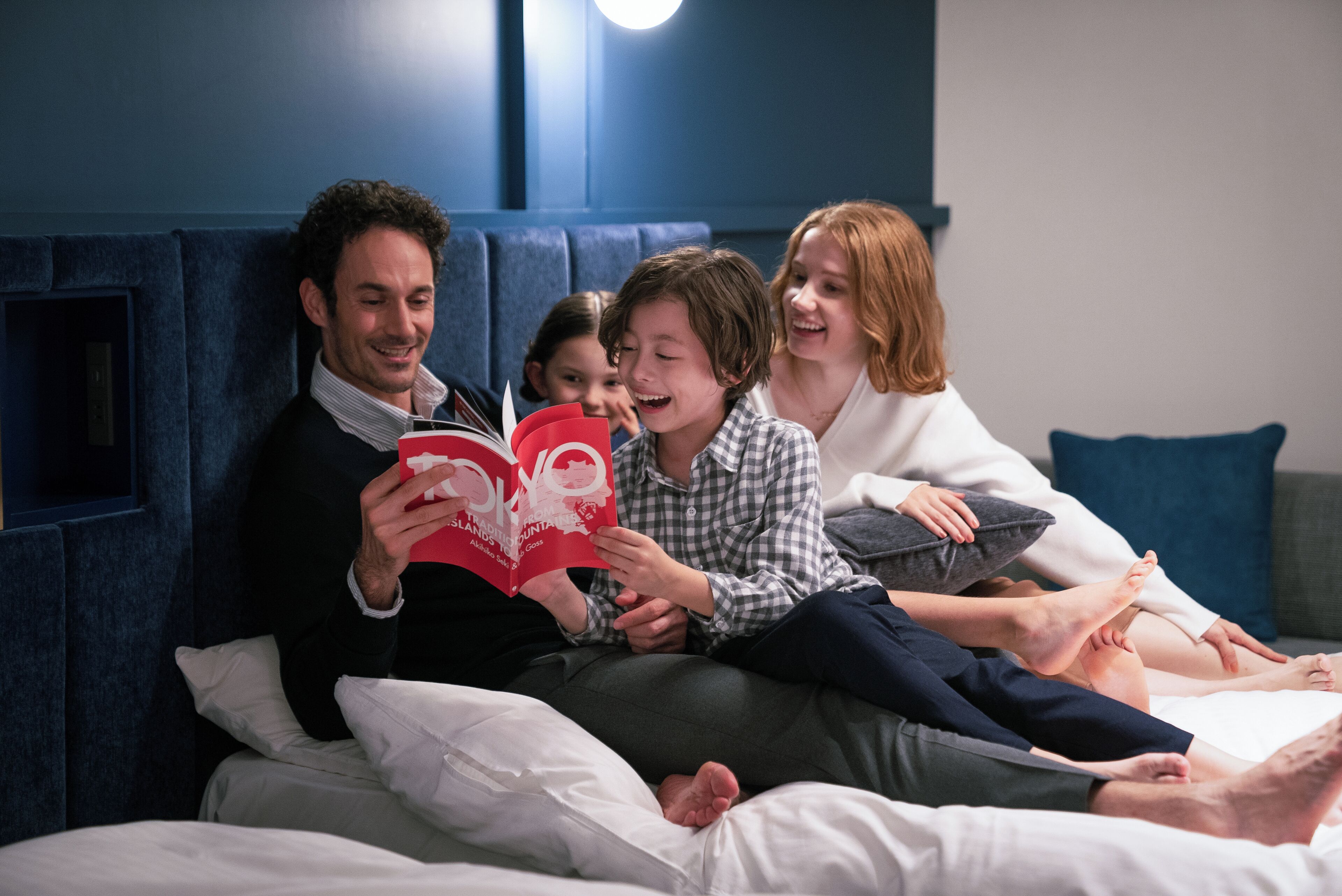 A family of four sitting on a bed in a room with a blue headboard and white bedding.