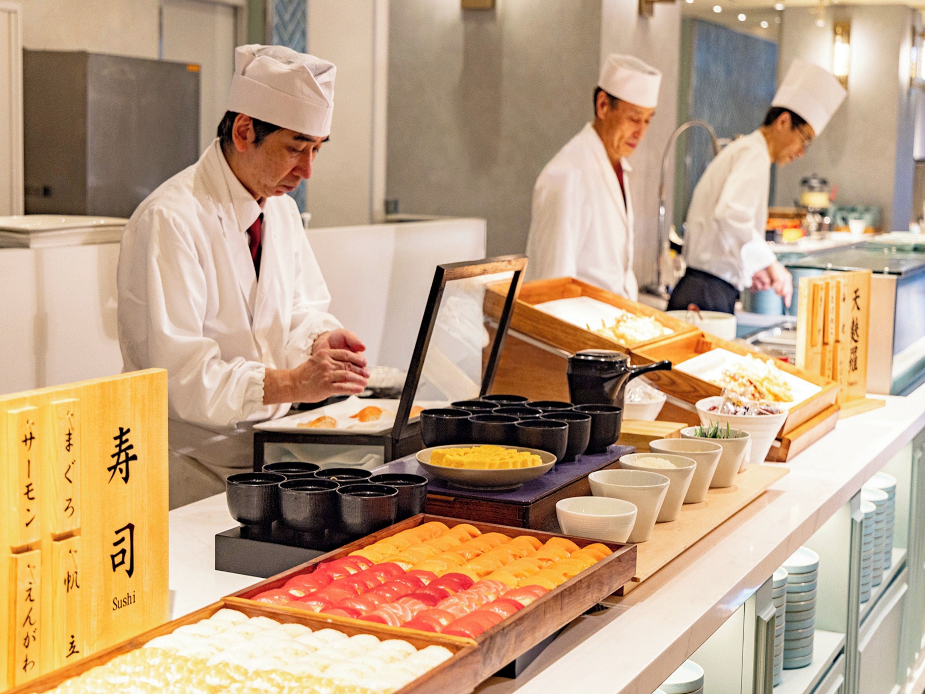 A sushi bar with chefs preparing food, a variety of sushi displayed on trays, and traditional Japanese writing on the side.