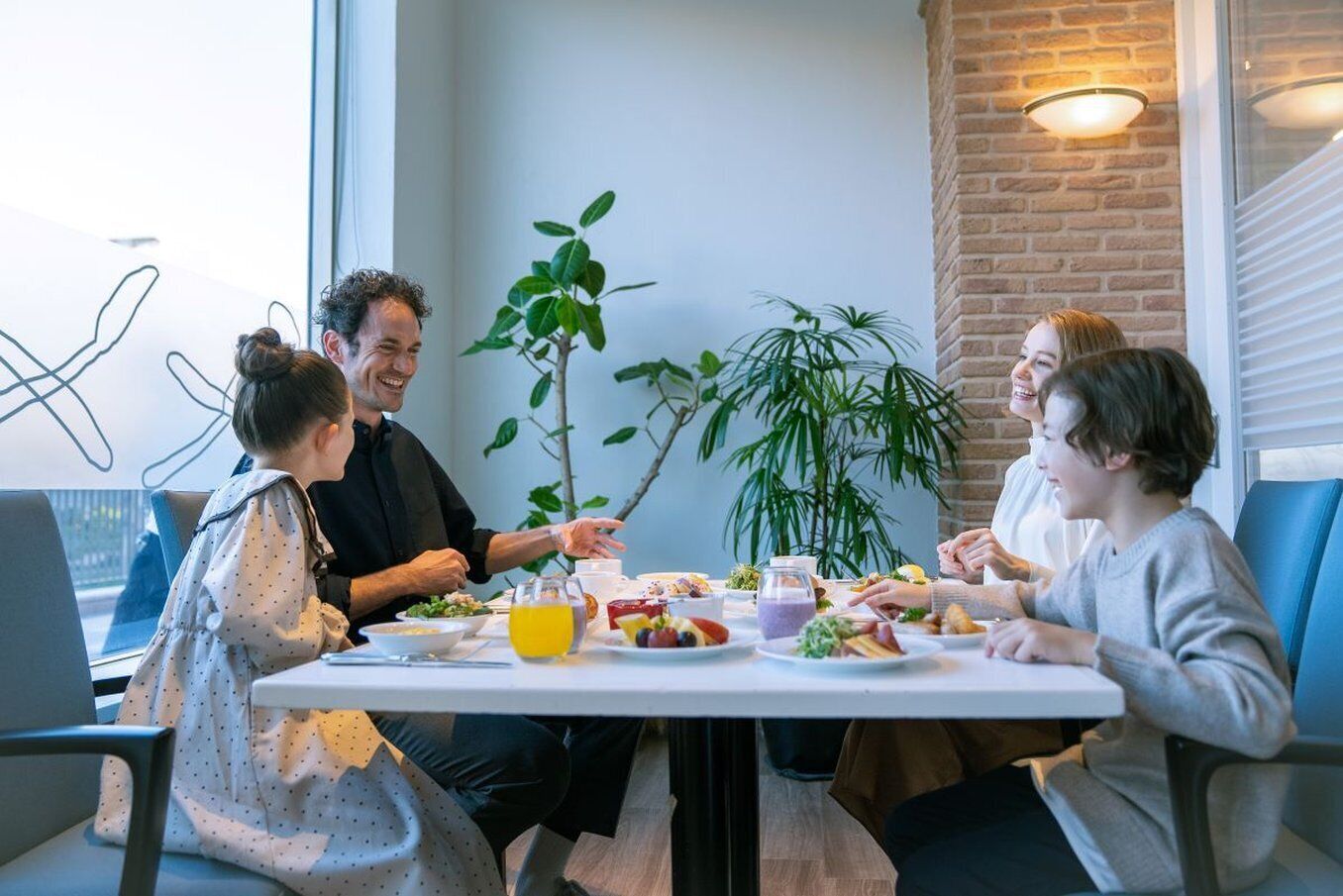 A group of people seated around a table with food and drinks, in a room with a brick wall and large windows.