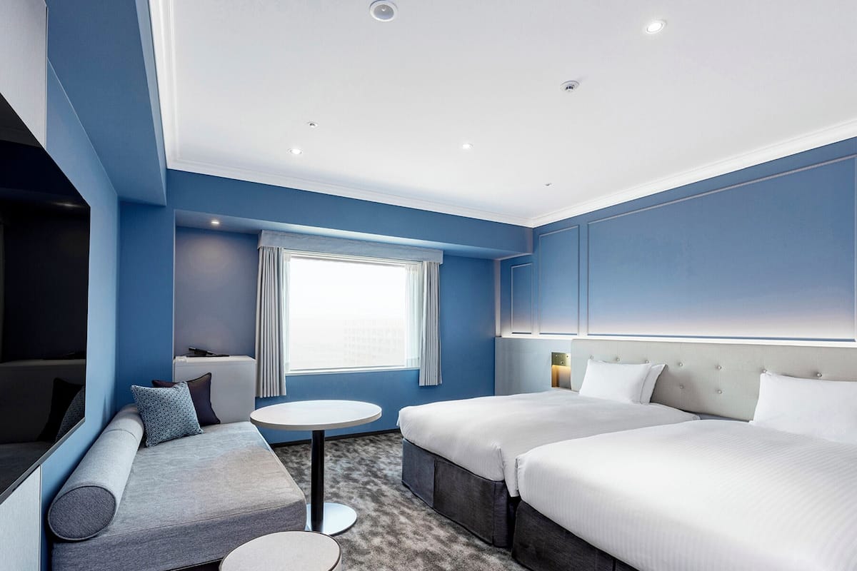 Moderni hotellihuone, jossa on sininen tehosteseinä, pyöreä pöytä, kaksi sänkyä, sohva ja televisio.