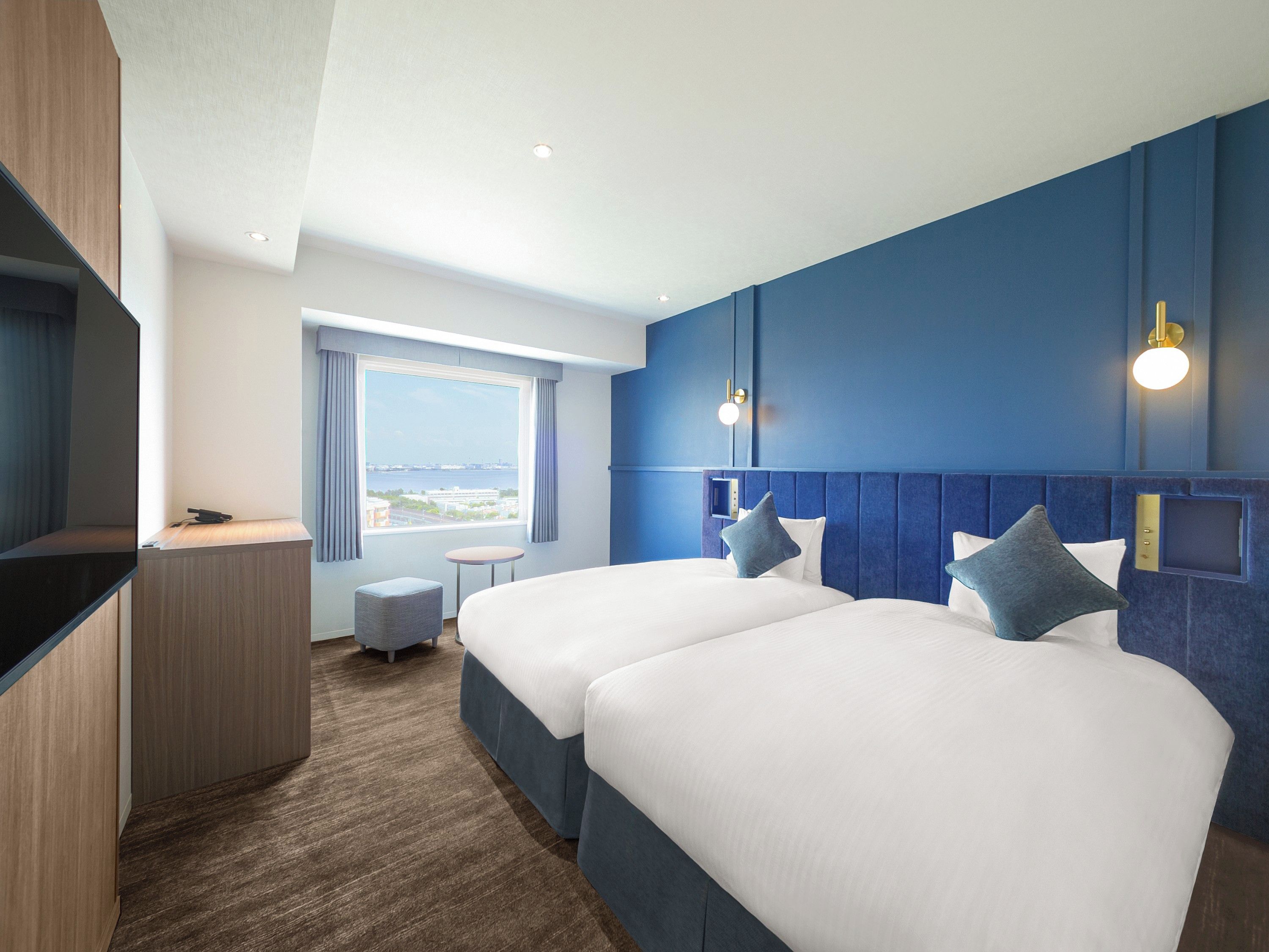 A hotel room with two beds, a desk, a chair, a window with a view, and wall-mounted lighting.