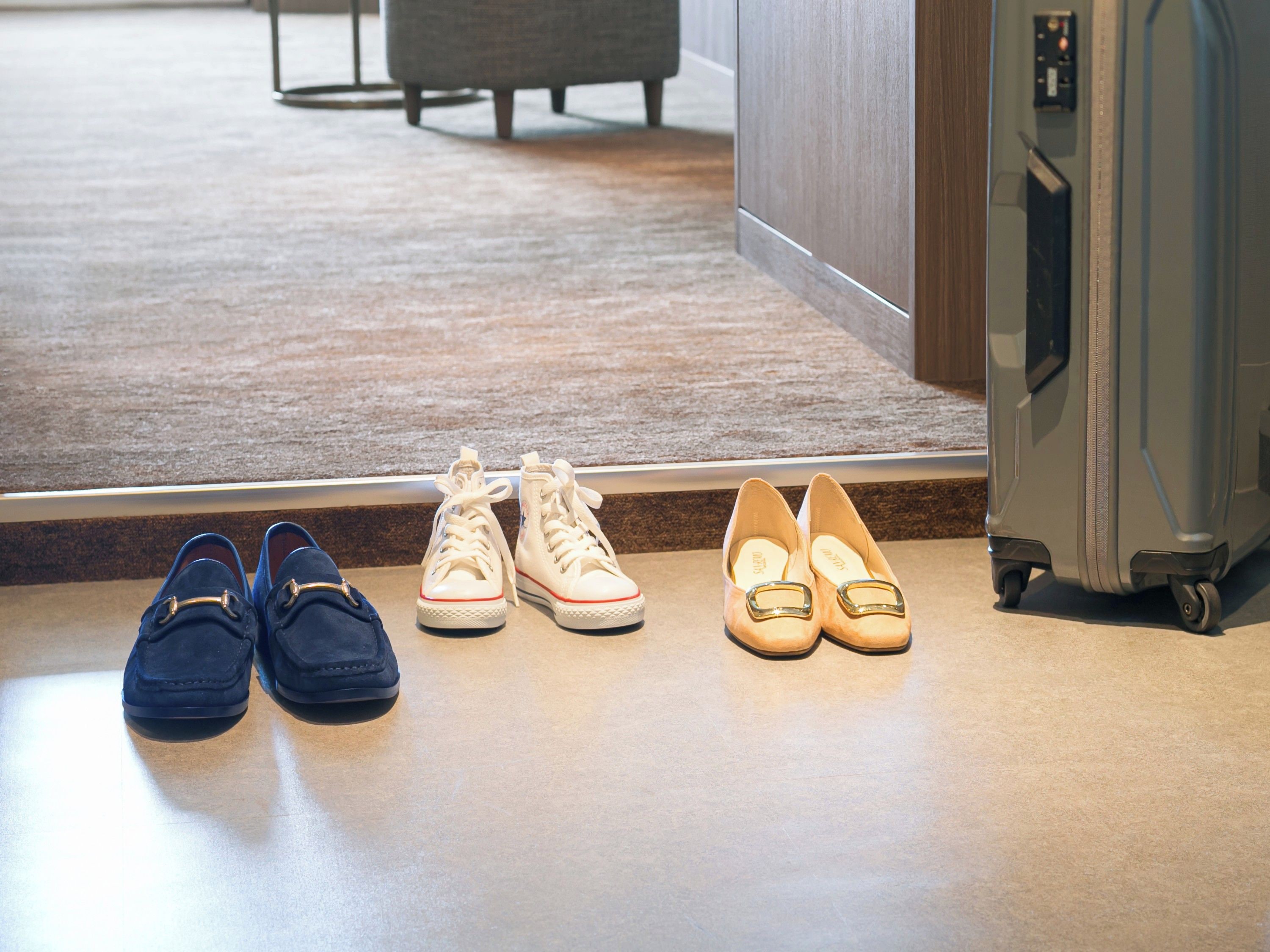 A pair of shoes on a carpeted floor near a door, with a suitcase and a piece of furniture in the background.