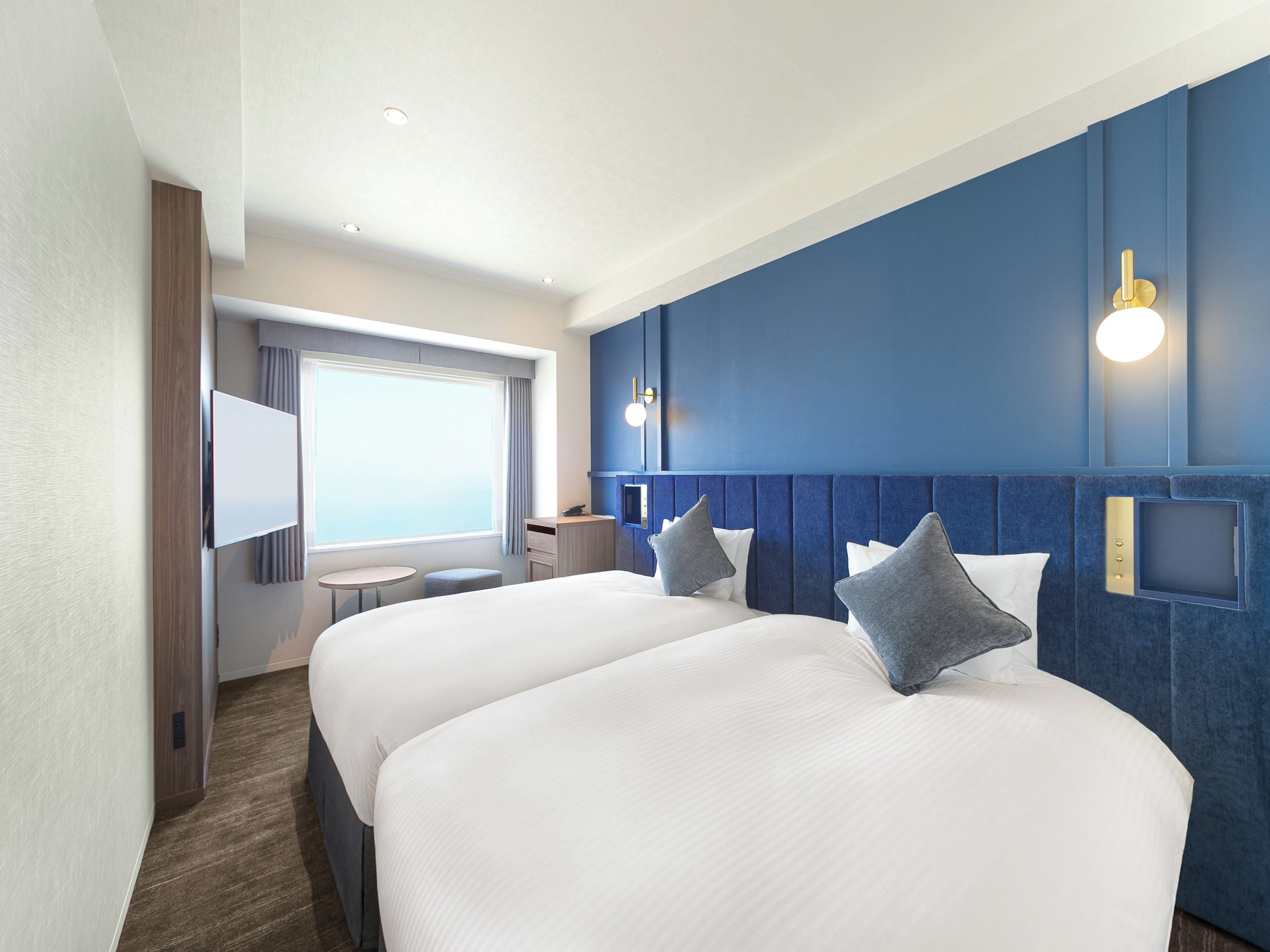 A hotel room with two beds, a desk, a chair, and a window.