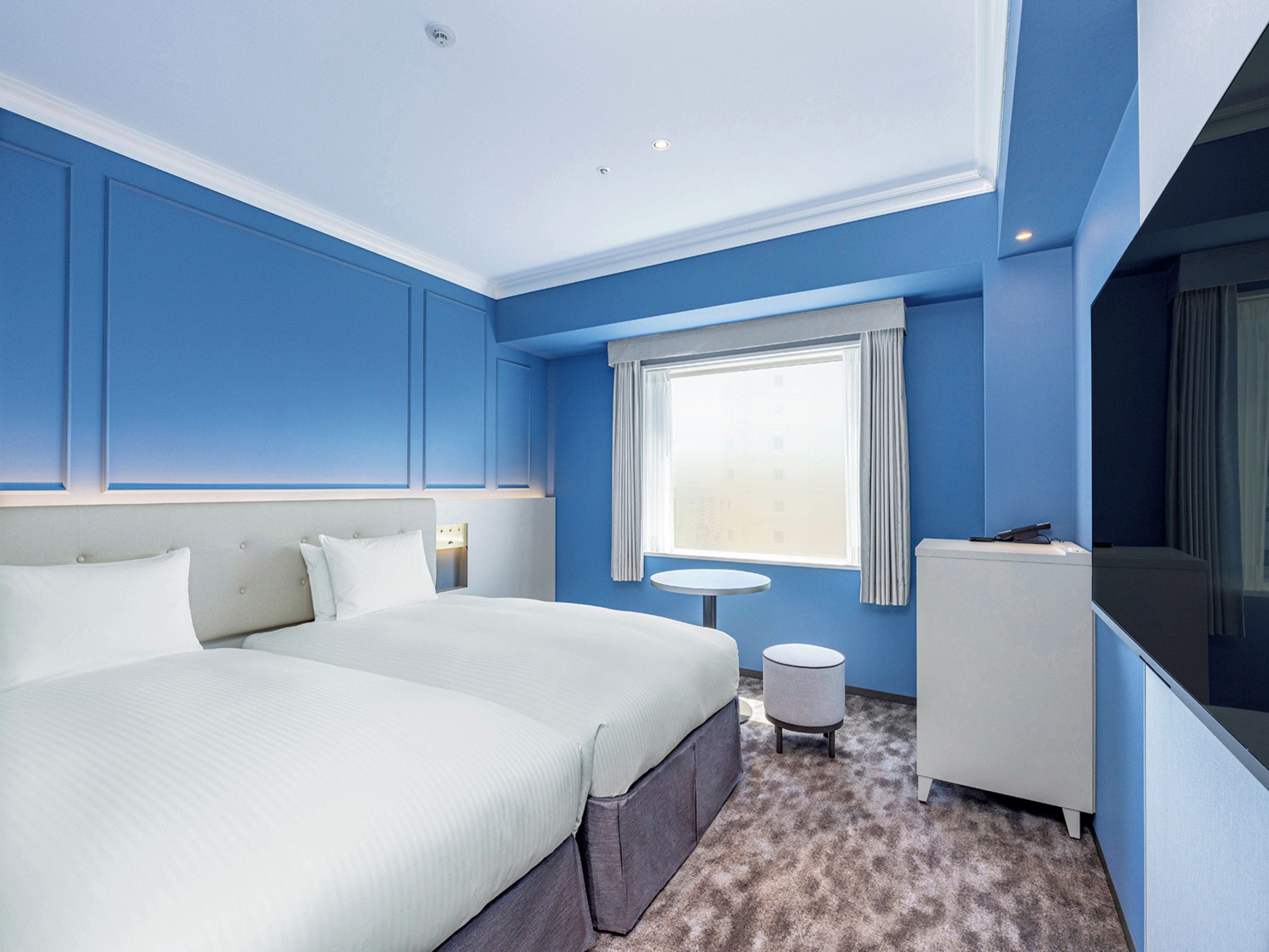 A hotel room with two beds, a small round table, a sofa, and a large window with curtains.