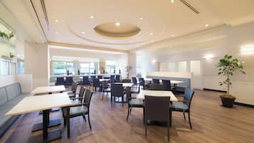 Un vaste espace de restauration intérieur, doté de nombreuses tables et chaises, d’un coin salon central et d’une grande fenêtre laissant entrer la lumière naturelle.