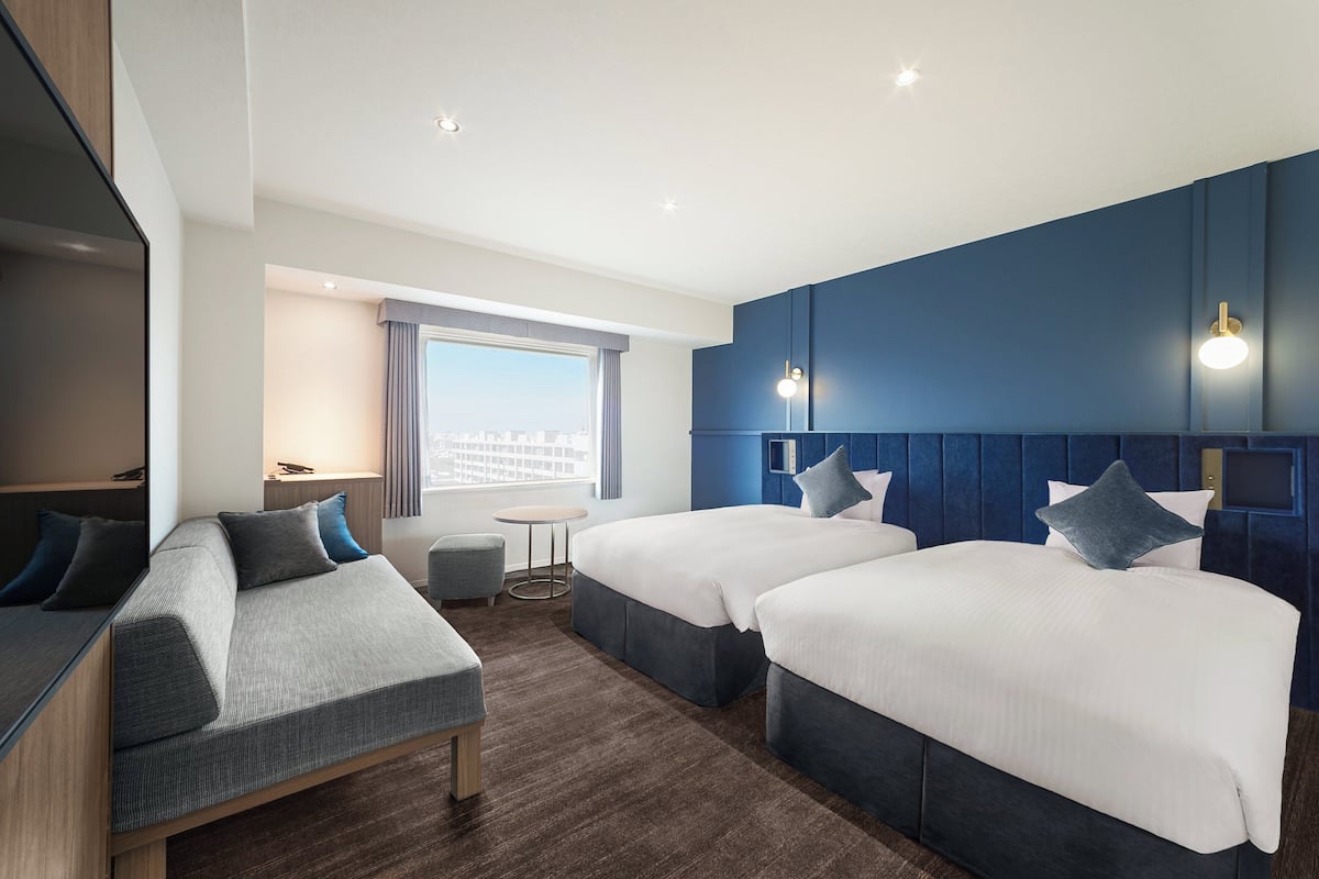 Hotellihuone, jossa on kaksi sänkyä, sohva, pieni pöytä ja näkymä kaupunkiin.