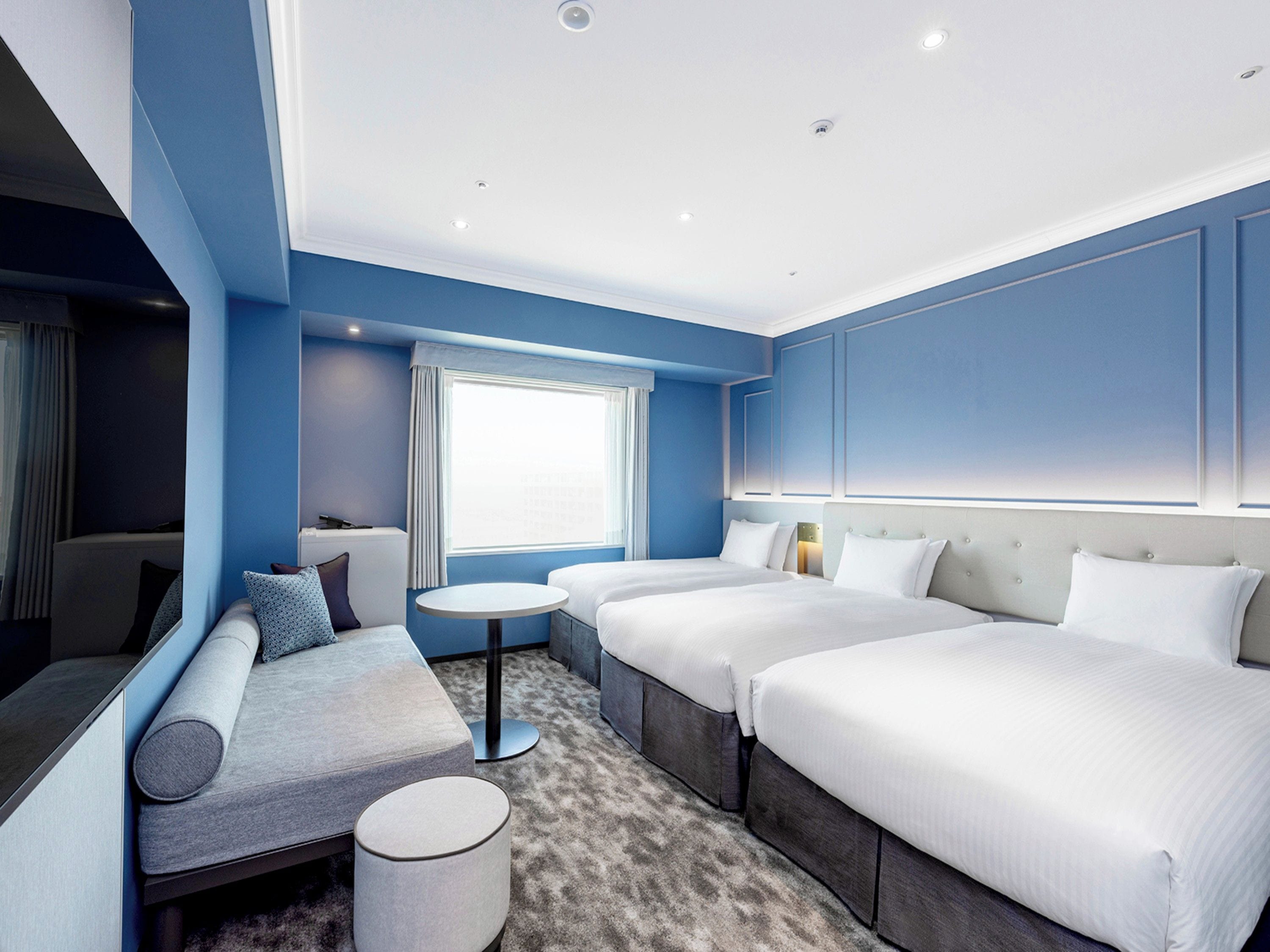 A hotel room with a sofa, two beds, a small table, and a window.