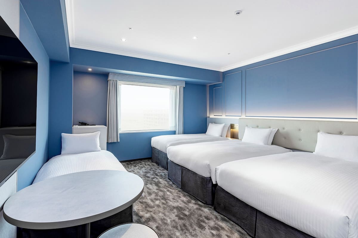 Hotellihuone, jossa on kaksi sänkyä, pieni pöytä, ikkuna verhoilla ja kattoon upotetut valaisimet.