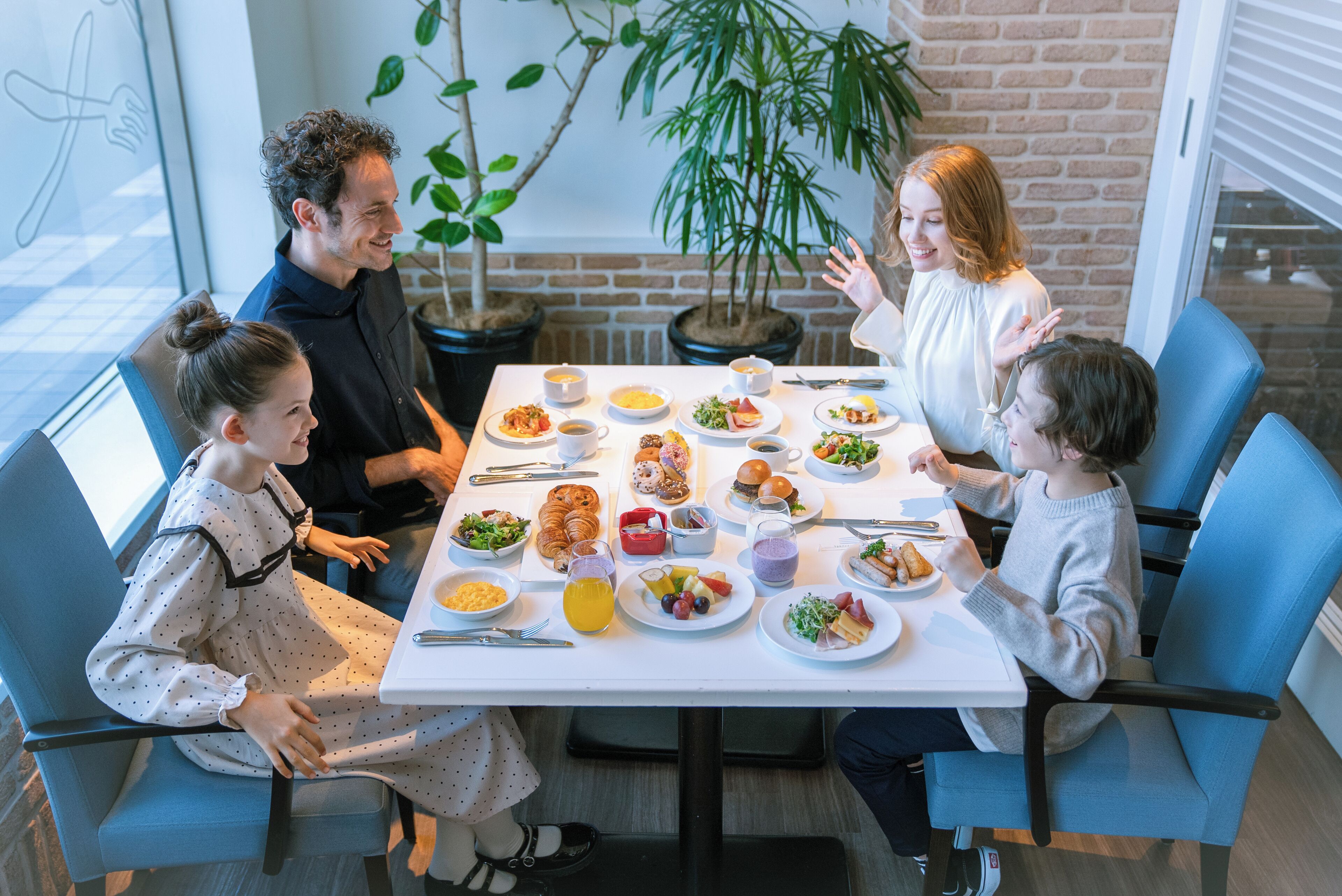 A group of people seated around a table with various dishes, in a room with brick walls and potted plants.