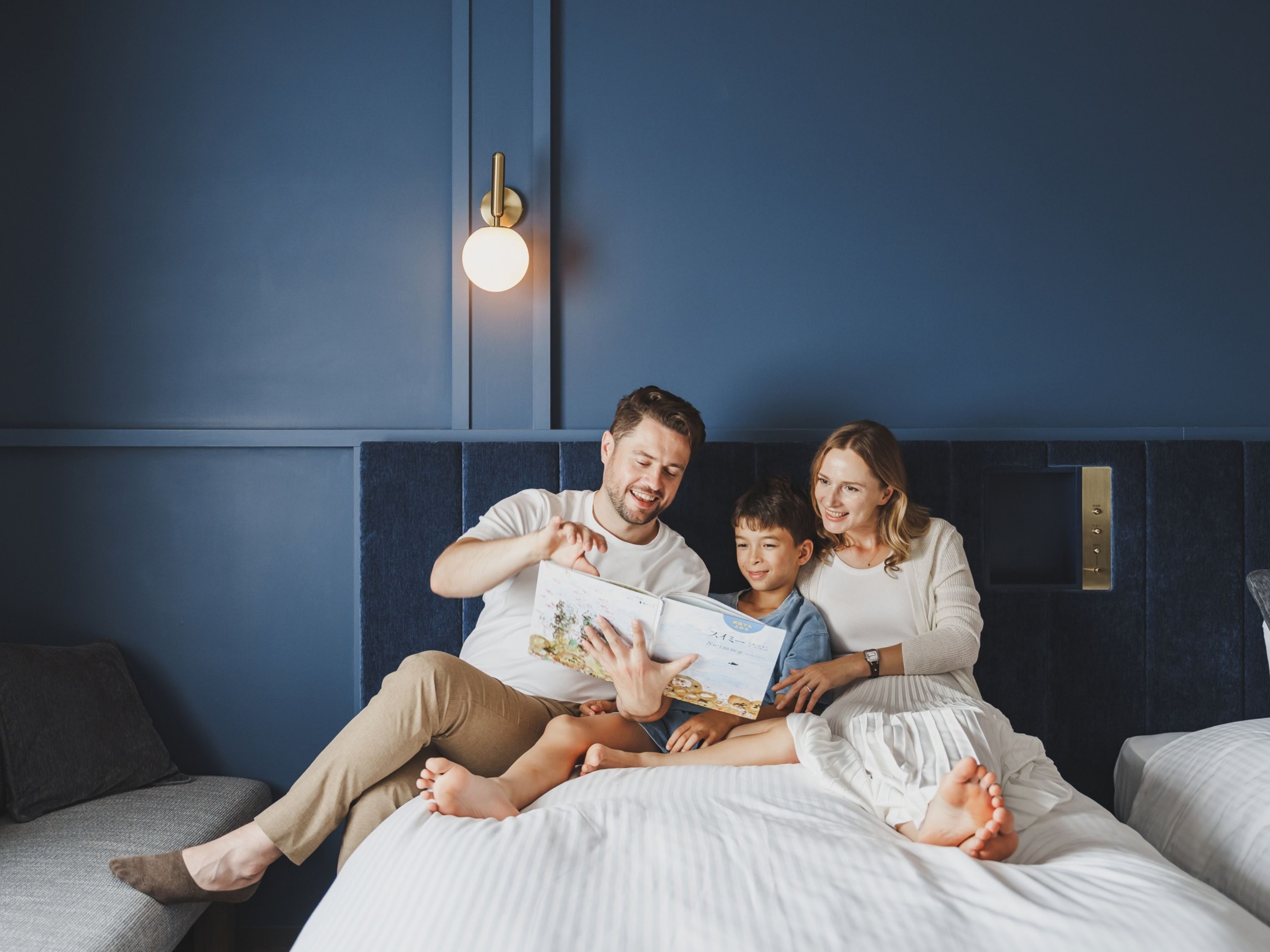 A family of three on a bed with a blue headboard, a wall-mounted light fixture, and a flat-screen TV.