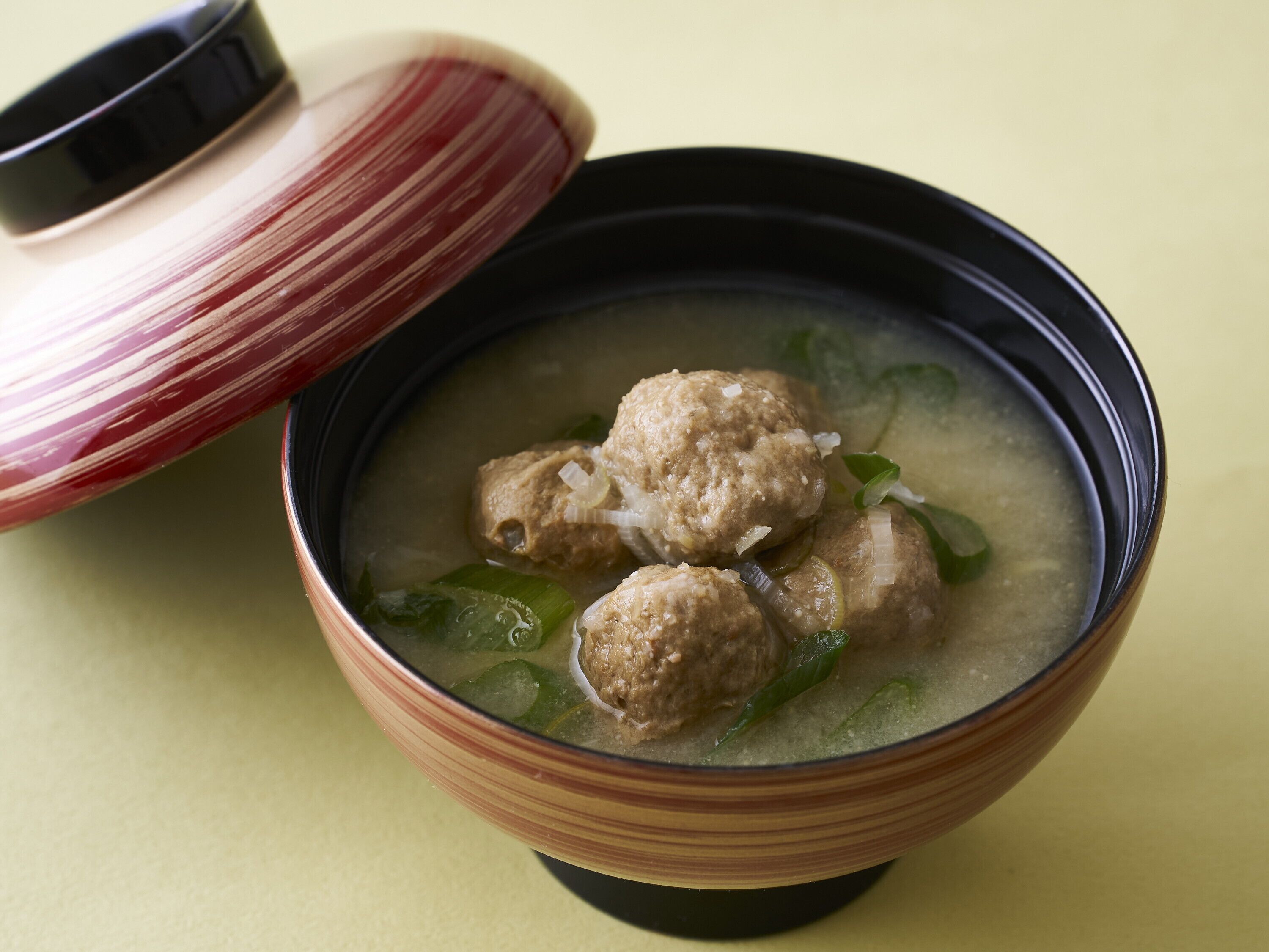 Un tazón de sopa con albóndigas y verduras verdes, acompañado de una tapa de cerámica a rayas.