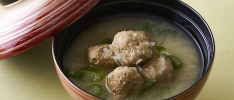 Un tazón de sopa con albóndigas y verduras verdes, acompañado de una tapa de cerámica a rayas.