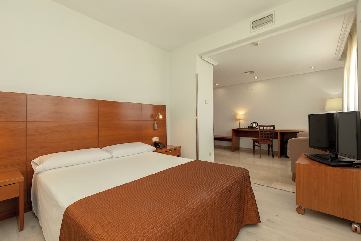 Quarto de hotel com cama grande, cabeceira de madeira, mesa de cabeceira e televisão sobre suporte de madeira.