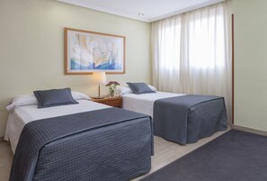 Ein Hotelzimmer mit zwei Betten, einem Nachttisch mit Lampe, einem gerahmten abstrakten Gemälde und einem großen Fenster mit Vorhängen.