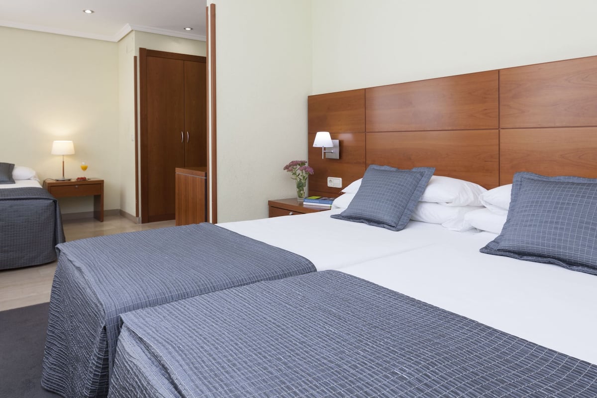 Quarto de hotel com duas camas, cabeceira de madeira, mesa de cabeceira com abajur e guarda-roupa.