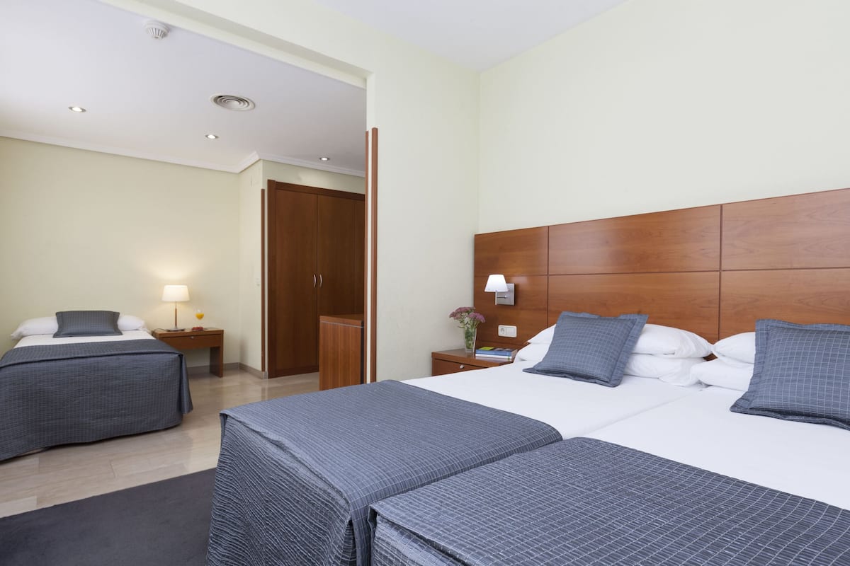 Quarto de hotel com duas camas, cabeceira de madeira, mesa de cabeceira com abajur e porta para outro cômodo.