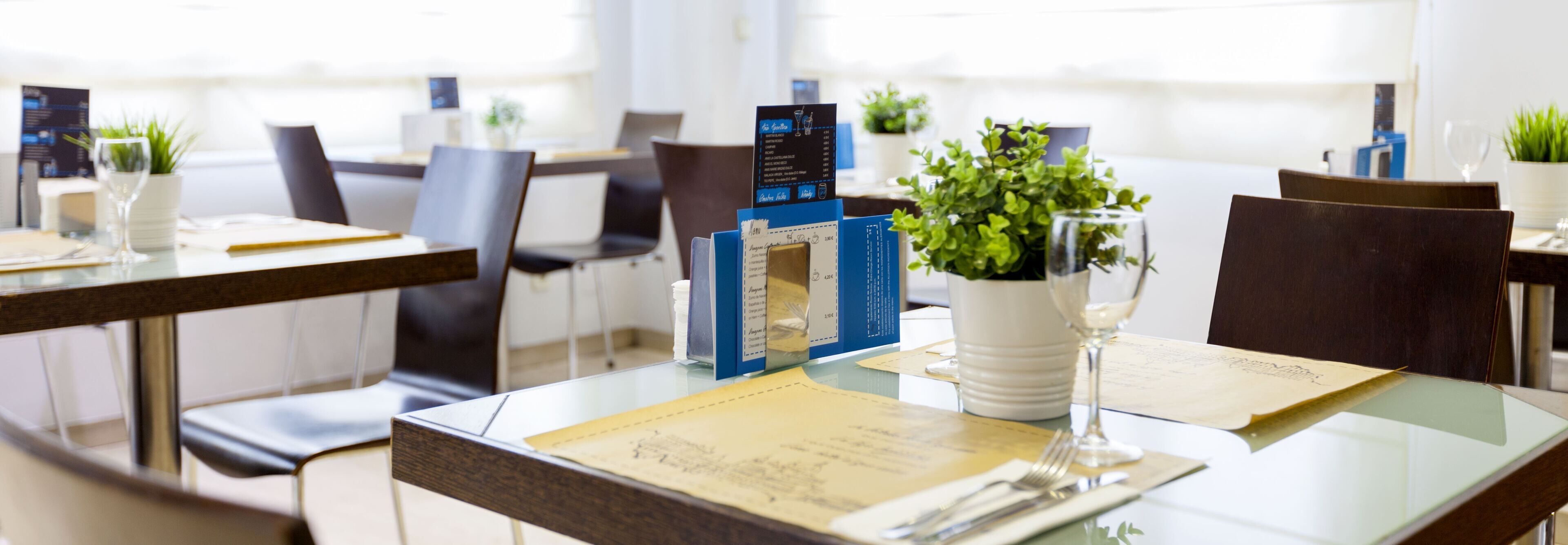 A restaurant setting with tables set for dining, menus, and a potted plant on the table.