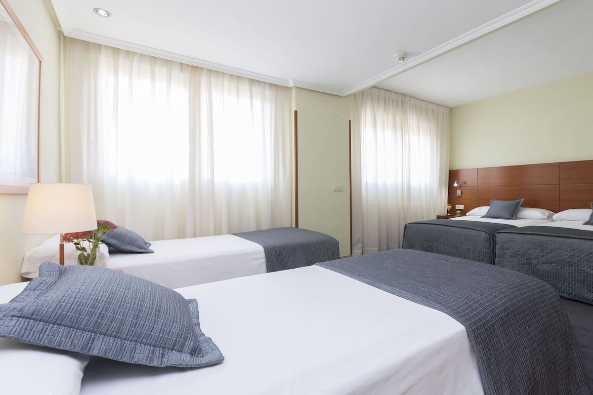Quarto de hotel com duas camas de solteiro, uma cama de casal, uma mesa de cabeceira com abajur e uma janela com cortinas.