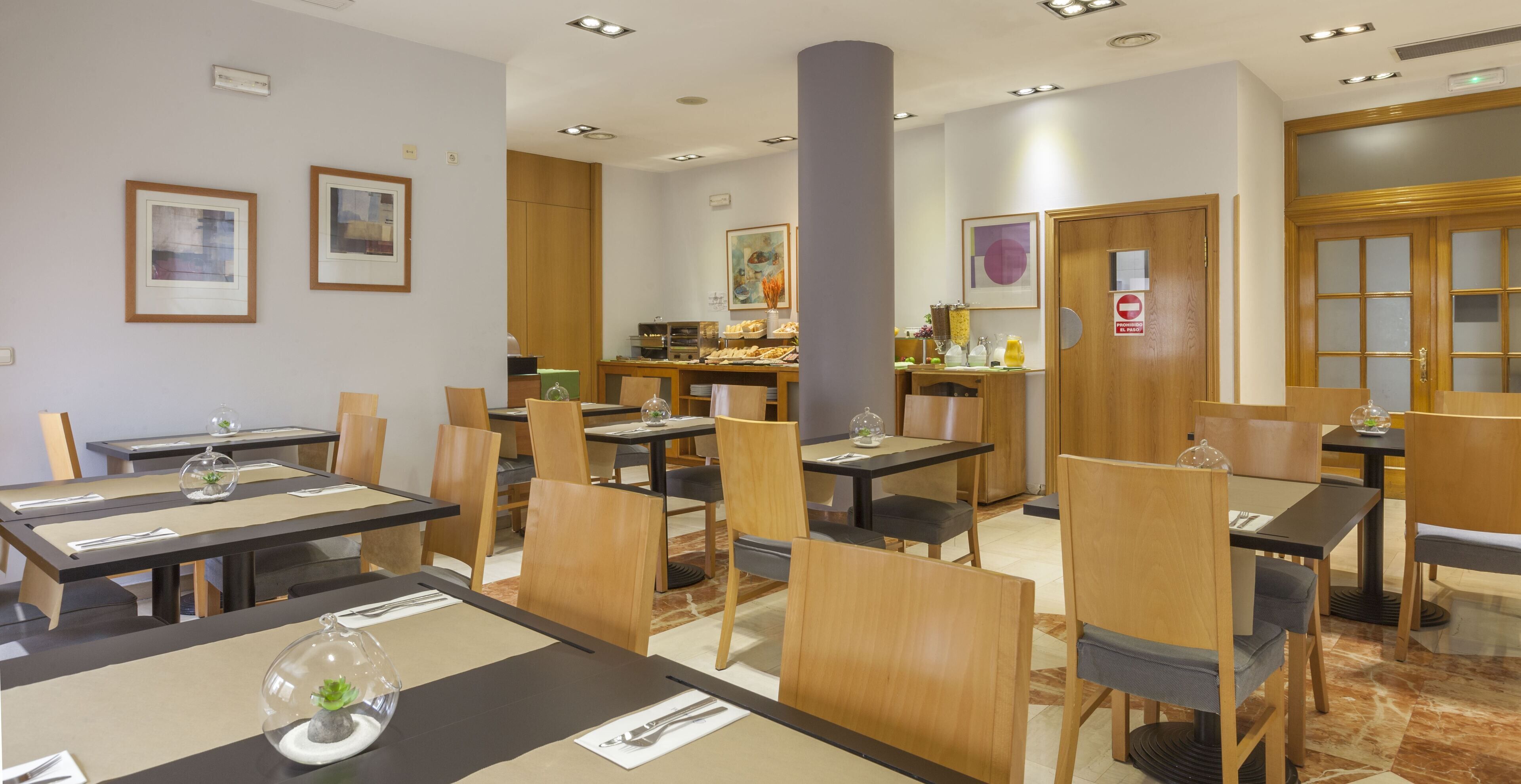 Un'area ristorazione in un hotel con diversi tavoli e sedie, un angolo caffè e opere d'arte appese alle pareti.