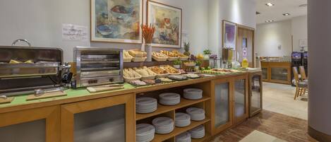 Zona de buffet con diversos alimentos, una máquina de café y un área de estar con sillas y una mesa.