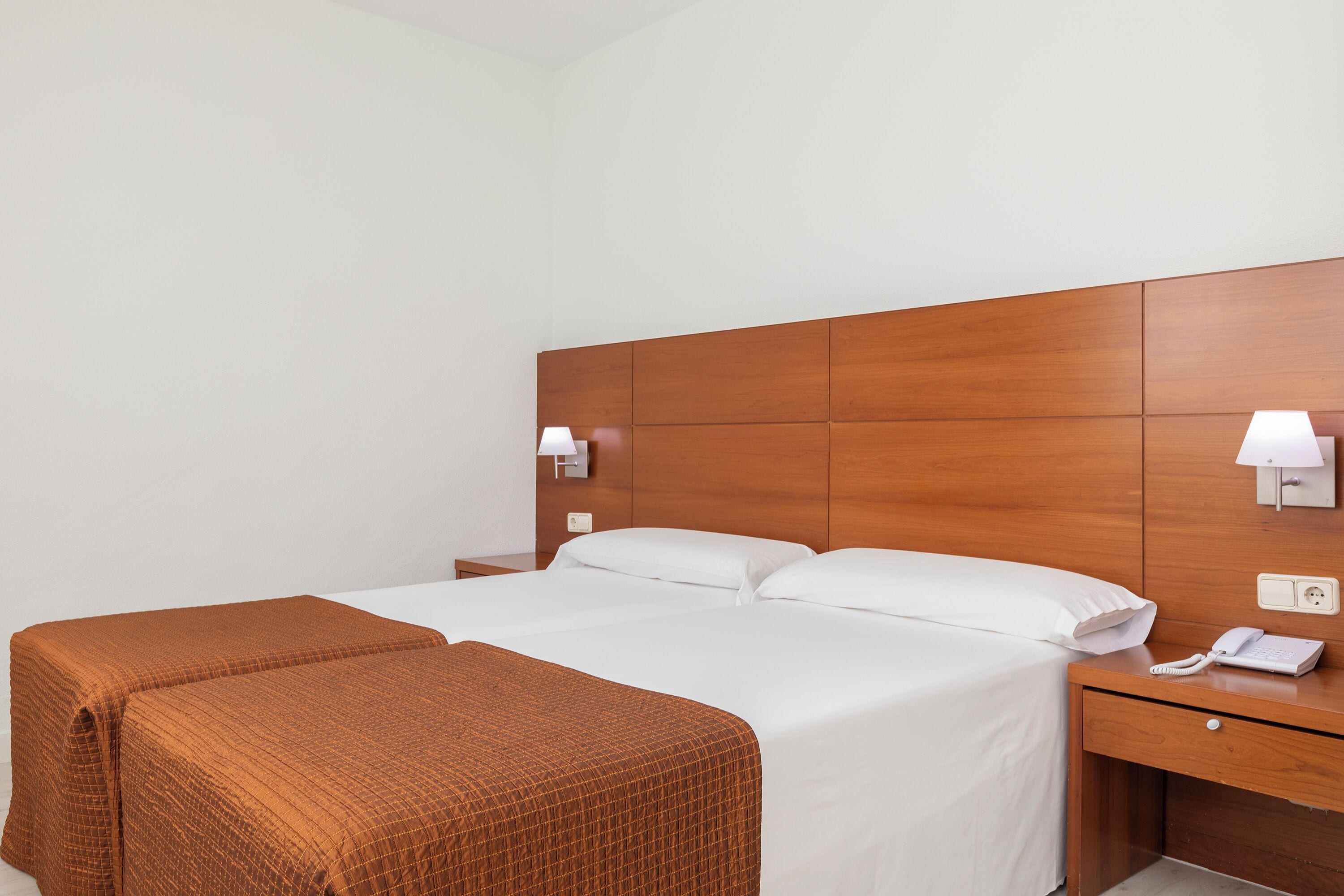 A hotel room with a wooden headboard, a bed with white linens, two bedside tables, and a telephone on one of the tables.