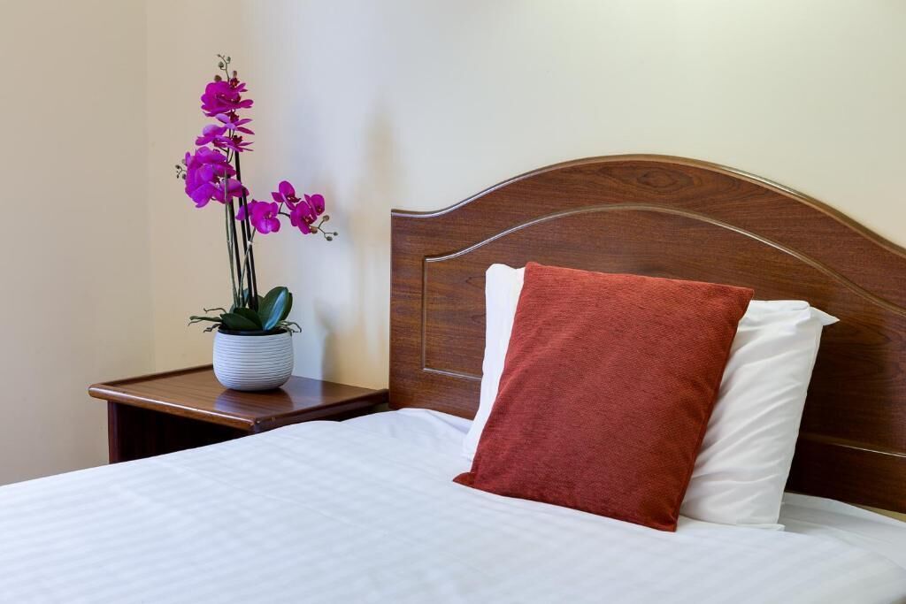 Una cama bien hecha con ropa blanca y un cojín marrón, un cabecero de madera y una mesita de noche con una planta en maceta.