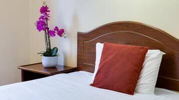 Una cama bien hecha con ropa blanca y un cojín marrón, un cabecero de madera y una mesita de noche con una planta en maceta.