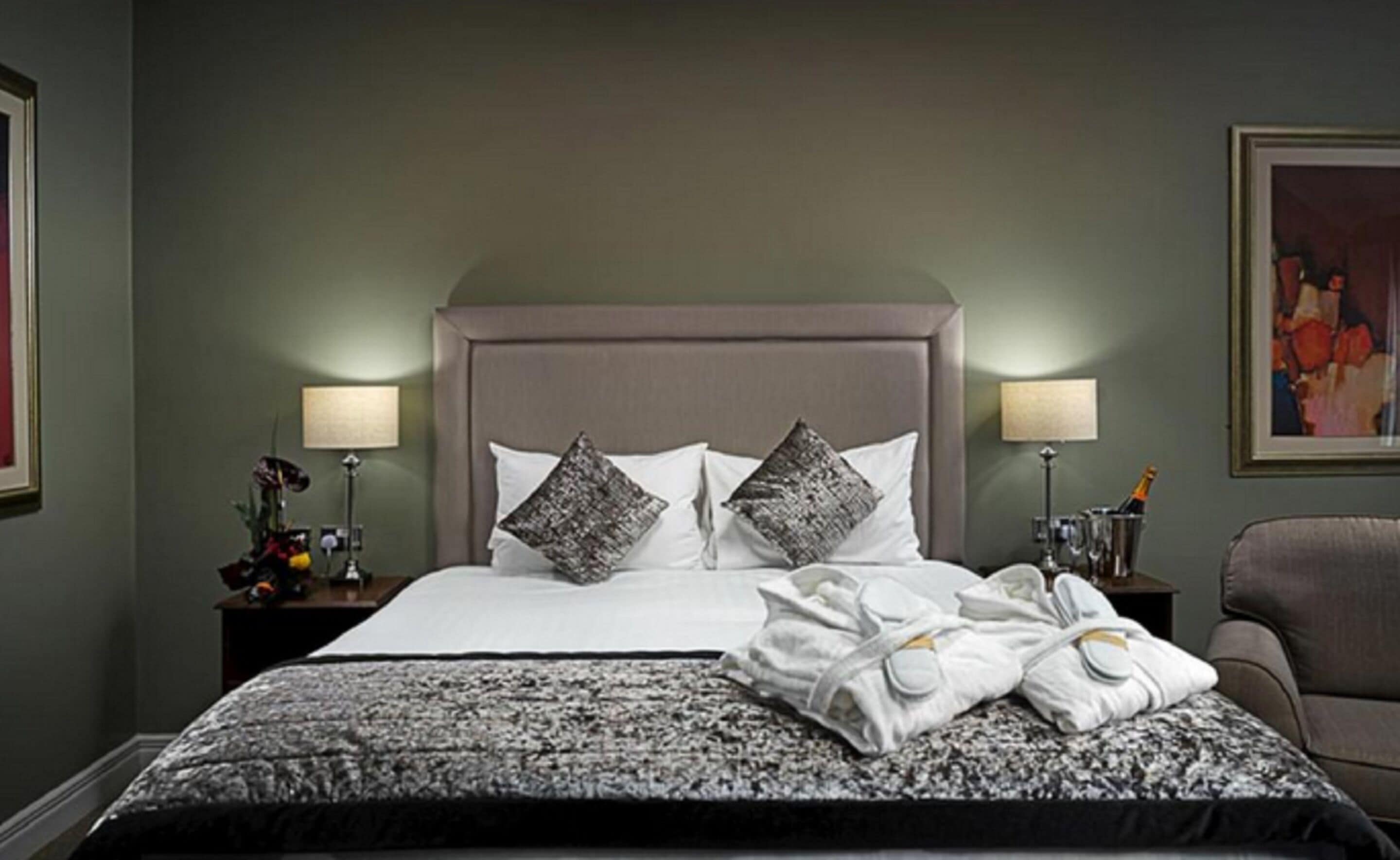 A hotel room with a large bed, two bedside lamps, a nightstand with a vase and a lamp, and a chair.